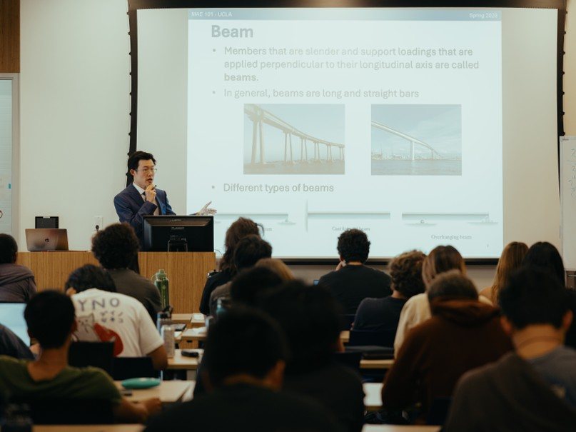 Yupeng Zhang lecturing to a full classroom at UCLA, with a slide on the types of beams projected behind him.