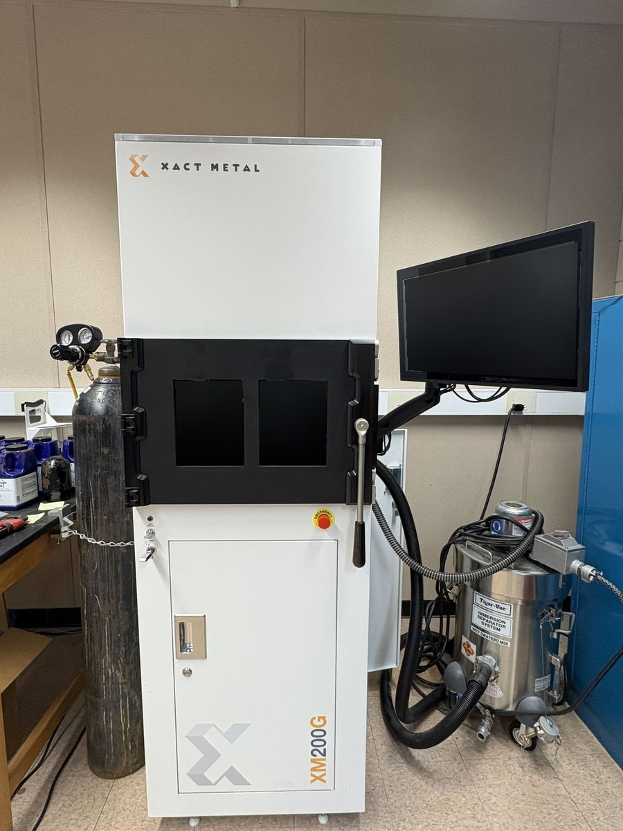 Xact Metal XM200G laser powder bed fusion printer in room E0040, with its nitrogen cylinder and regulator at left, a monitor on an articulated arm at right, and a Tiger-Vac immersion separator vacuum connected by hose.